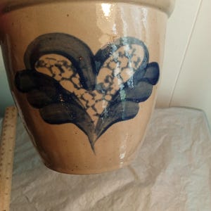 May include: A tan ceramic planter with a blue and white floral design. The planter has a wide rim and a slightly tapered body. The floral design is centered on the front of the planter and is made up of blue petals around a white and blue center.