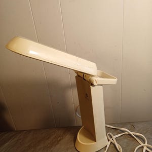 May include: A cream-colored desk lamp with a rectangular base and a long, adjustable shade. The lamp has a handle on the back and a white cord. The lamp is made of plastic and is designed for desk use.