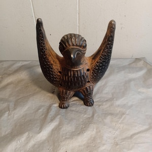 Può includere: Una scultura di uccello in ceramica marrone scuro con le ali spiegate. L'uccello presenta dettagliate decorazioni di piume e una superficie strutturata. La scultura è in piedi su tre piccoli piedi e ha un aspetto invecchiato.