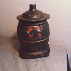 Puede incluir: Un tarro de galletas de cerámica negra con forma de estufa vintage. El tarro tiene un cuerpo redondo con tapa y un panel decorativo con diseños florales naranjas y amarillos. La base tiene un asa.