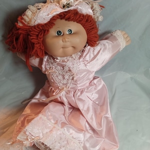 Può includere: Una bambola con capelli in lana color mogano, occhi verdi e viso rotondo. La bambola indossa un abito in raso rosa chiaro con bordi in pizzo bianco, un corpetto in pizzo sovrapposto e piccoli fiocchi di nastro. Una cuffietta rosa abbinata.