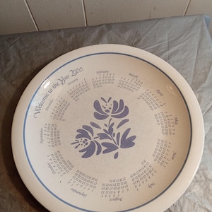 May include: A white ceramic plate with a blue floral design in the center and a calendar for the year 2000 around the rim. The plate has the text "Welcome to the Year 2000" in a script font.
