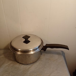 May include: Stainless steel saucepan with a lid and a long, dark brown handle. The lid has a black, diamond-shaped handle. The pot is sitting on a light gray surface, with a white wall in the background.