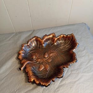 May include: A decorative leaf-shaped dish with a bronze finish. The dish features detailed leaf and acorn designs, with a darker interior and lighter bronze edges. The dish is likely made of metal and is suitable for decorative purposes.