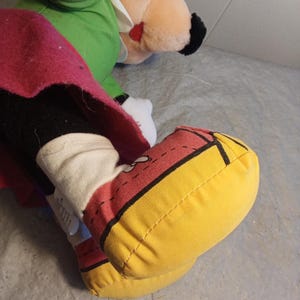 May include: Close-up of a plush toy foot in yellow, red, and black. The foot is part of a larger plush toy, with a white sock and a red and black shoe design. Another plush toy is visible in the background.