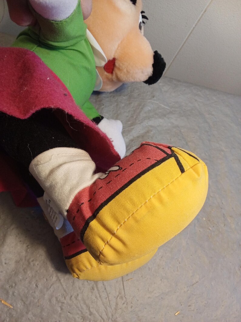 May include: Close-up of a plush toy foot in yellow, red, and black. The foot is part of a larger plush toy, with a white sock and a red and black shoe design. Another plush toy is visible in the background.