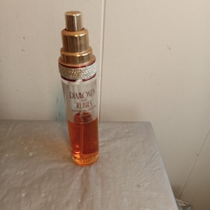 May include: A perfume bottle with a gold-colored cap and a clear glass body. The bottle is filled with an orange-colored liquid. The text "Diamonds and Rubies" is visible on the bottle.