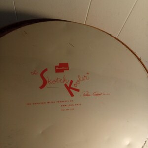 May include: Vintage "Skotch Kooler" metal cooler lid. The lid is off-white with red lettering and a red square logo. The text includes "the Skotch Kooler" and "The Hamilton Metal Products Co., Hamilton, Ohio."