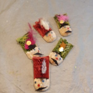 Puede incluir: Cinco figuras decorativas hechas a mano que se asemejan a soldados de juguete. Cada figura tiene un sombrero rojo o verde, una cara pintada con bigote negro y un adorno de pluma o flor. Las figuras están dispuestas sobre una superficie gris claro.