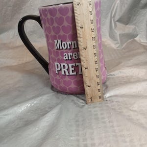 Puede incluir: Una taza de cer&aacute;mica rosa con asa negra. La taza est&aacute; decorada con peque&ntilde;os corazones rosas y las palabras "Mornings are PRETTY" en blanco. La taza mide aproximadamente 12.7 cm de alto.
