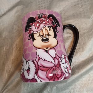 Puede incluir: Taza de cer&aacute;mica rosa con un personaje de dibujos animados con una bata y antifaz rosas. La taza tiene un asa negra y un patr&oacute;n de corazones rosas. El personaje es un rat&oacute;n con orejas negras y un lazo.