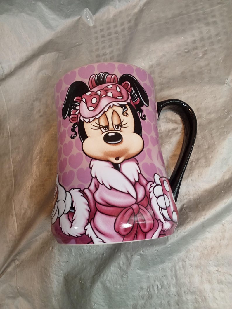 Puede incluir: Taza de cer&aacute;mica rosa con un personaje de dibujos animados con una bata y antifaz rosas. La taza tiene un asa negra y un patr&oacute;n de corazones rosas. El personaje es un rat&oacute;n con orejas negras y un lazo.