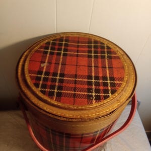 May include: A vintage round tin container with a red and black plaid pattern on the lid and body. The container has a light brown trim and a pink handle. The container is likely used for storage.