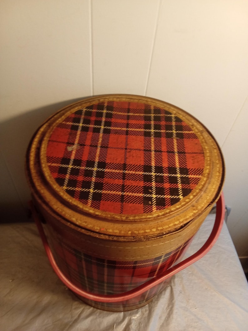 May include: A vintage round tin container with a red and black plaid pattern on the lid and body. The container has a light brown trim and a pink handle. The container is likely used for storage.