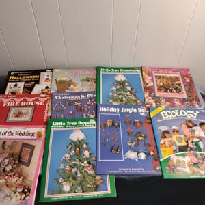 Könnte beinhalten: Eine Sammlung von Bastelbüchern mit bunten Umschlägen zu verschiedenen Themen, darunter Halloween, Weihnachten und Ökologie. Die Bücher tragen Titel wie "Halloween Iron-on Transfer Book", "Kleine Baumschmuck", "Weihnachten 1968", "Weihnachtsglocken" und "Ökologie".