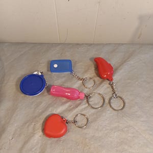 Puede incluir: Una colección de llaveros coloridos de varias formas, incluyendo un círculo azul, un rectángulo azul, una botella rosa, un corazón rojo y una lágrima roja. Cada llavero tiene un anillo de metal plateado.