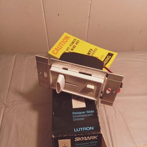 Puede incluir: Una caja negra con un interruptor de atenuación incandescente Lutron Skylark Designer Slide blanco. La caja tiene el número de modelo S-603P impreso en ella. El interruptor está conectado a una placa de pared blanca con un cable negro que sale de él. El interruptor está sobre una superficie de madera con una regla al lado.