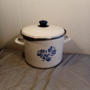 Puede incluir: Olla de esmalte blanco con tapa y dos asas. La olla presenta un diseño floral azul y un pomo azul oscuro en la tapa. Está sobre una superficie gris. Adecuada para cocinar.