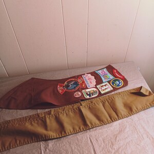 May include: A brown sash with several colorful patches and a tan belt, likely for a youth organization. The patches feature various designs and text, including the word "Maine". The belt is a solid tan color.