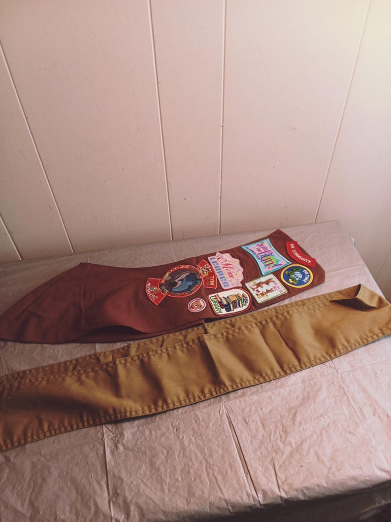 May include: A brown sash with several colorful patches and a tan belt, likely for a youth organization. The patches feature various designs and text, including the word "Maine". The belt is a solid tan color.