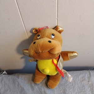 Puede incluir: Un hipopótamo de peluche marrón que lleva una chaqueta dorada con una cinta roja. El hipopótamo tiene pelo amarillo en el vientre y un lazo rojo en la cabeza.