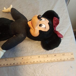 May include: A vintage Minnie Mouse plush doll. The doll has a black body, ears, and a red bow. The face is tan with black eyes and a black nose. A wooden ruler is next to the doll, measuring up to 22 inches.