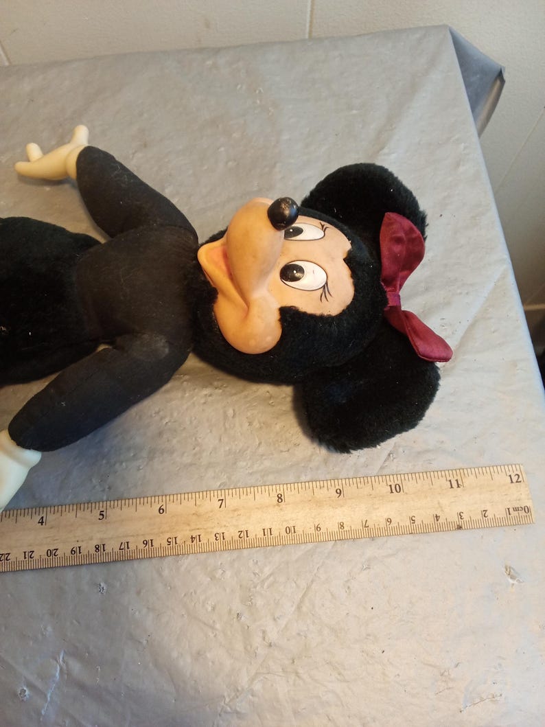 May include: A vintage Minnie Mouse plush doll. The doll has a black body, ears, and a red bow. The face is tan with black eyes and a black nose. A wooden ruler is next to the doll, measuring up to 22 inches.