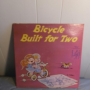 May include: A vintage game box cover with a pink background and the title "Bicycle Built for Two" in yellow letters. The cover features an illustration of two children riding a tricycle, a dog running in the background, and a drawing of a bicycle on a piece of paper.