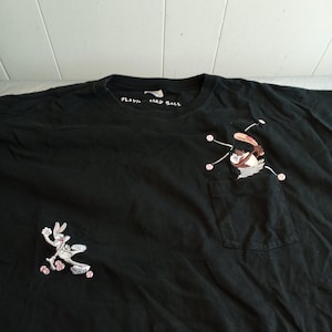May include: Black t-shirt with Looney Tunes baseball-themed graphics. Features Bugs Bunny running and Wile E. Coyote batting. The text "PLAYING HARD BALL" is printed on the neckline. Includes a pocket on the chest.
