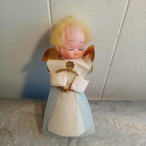 May include: A vintage angel Christmas ornament. The angel has a painted face with closed eyes, blonde hair, and gold wings. It wears a white dress with a light blue tulle skirt and a gold star-shaped embellishment.
