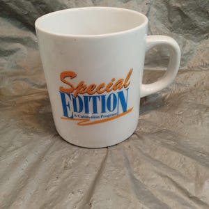 Puede incluir: Una taza de cerámica blanca con el texto Special EDITION A Cablevision Program impreso en el frente. La palabra Special está en letra cursiva naranja, mientras que EDITION está en letras mayúsculas azules, subrayada por una línea naranja.