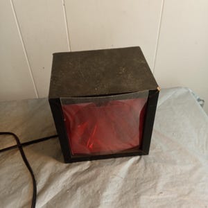 May include: A black, square box with a red interior. The box has a dark, textured top. A black electrical cord extends from the side. The box is on a white surface.