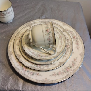 Puede incluir: Un juego de cuatro platos de cena de porcelana blanca con un patrón floral y ribete dorado. Los platos están apilados uno encima del otro, con una taza de té y un platillo a juego en la parte superior.