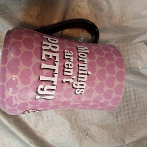 Puede incluir: Una taza de cer&aacute;mica rosa con asa negra. La taza est&aacute; cubierta con un patr&oacute;n de peque&ntilde;os corazones rosas. Las palabras "Mornings aren't PRETTY!" est&aacute;n impresas en blanco en el lateral.