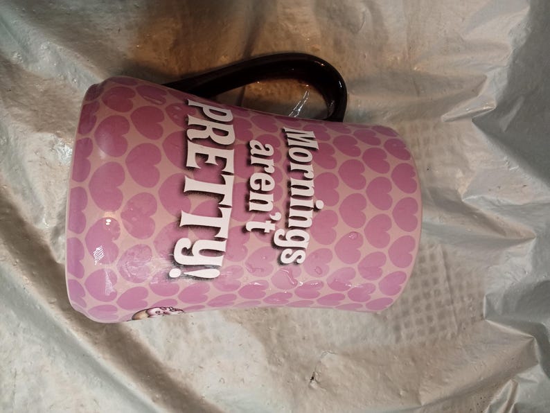 Puede incluir: Una taza de cer&aacute;mica rosa con asa negra. La taza est&aacute; cubierta con un patr&oacute;n de peque&ntilde;os corazones rosas. Las palabras "Mornings aren't PRETTY!" est&aacute;n impresas en blanco en el lateral.