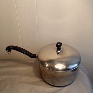 Può includere: Una casseruola in acciaio inossidabile con coperchio e manico nero. Il manico ha un anello all'estremità. Il coperchio ha un pomello nero. La pentola è su una superficie chiara.