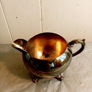 Op de afbeelding: Een zilverkleurige, ronde creamer met een tuit en handvat. De binnenkant heeft een gouden tint. De creamer staat op drie decoratieve voetjes. Het ontwerp is vintage en elegant.