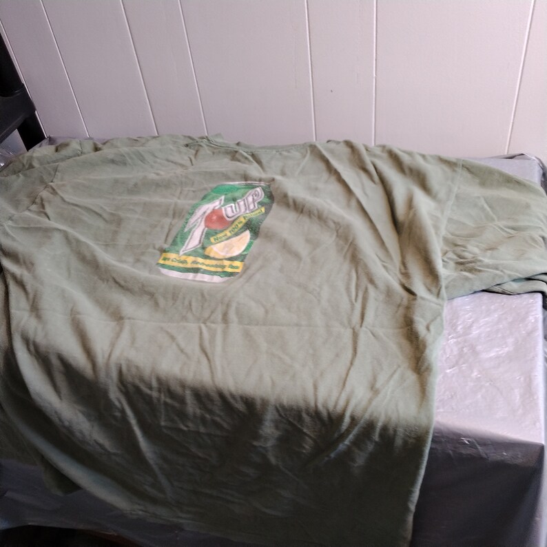 Puede incluir: Camiseta verde con un logotipo vintage de 7 Up que presenta un lim&oacute;n y el texto "7 Up, Now with a Twist!" y "The Only Drink Refreshingly You!"