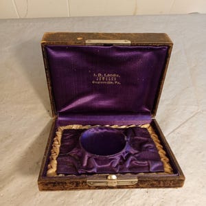 Puede incluir: Caja de joyería antigua abierta con exterior marrón oscuro e interior de terciopelo morado. La caja tiene un cierre y bisagras doradas. En el interior se lee el texto "I. D. Landis, JEWELER, Coatesville, Pa."