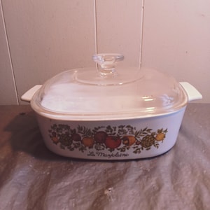 May include: White ceramic casserole dish with a glass lid. The dish has a floral design with fruits and vegetables. The text "Le Marseillaise" is printed on the bottom of the dish.