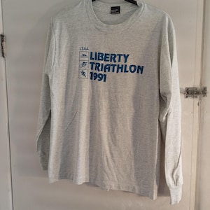 Puede incluir: Una camiseta de manga larga gris jaspeado con el texto "LIBERTY TRIATHLON 1991" en azul. La camiseta presenta tres pequeños gráficos azules encima del texto, que representan la natación, el ciclismo y la carrera. La camiseta está colgada en una percha negra.