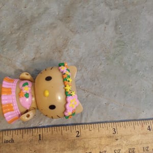 Op de afbeelding: Een bruin plastic beeldje van een kat met zwarte ogen en een gele neus. De kat draagt een roze en oranje outfit en een bloemenhoofdband. Een houten liniaal met inch-metingen ligt naast het beeldje.