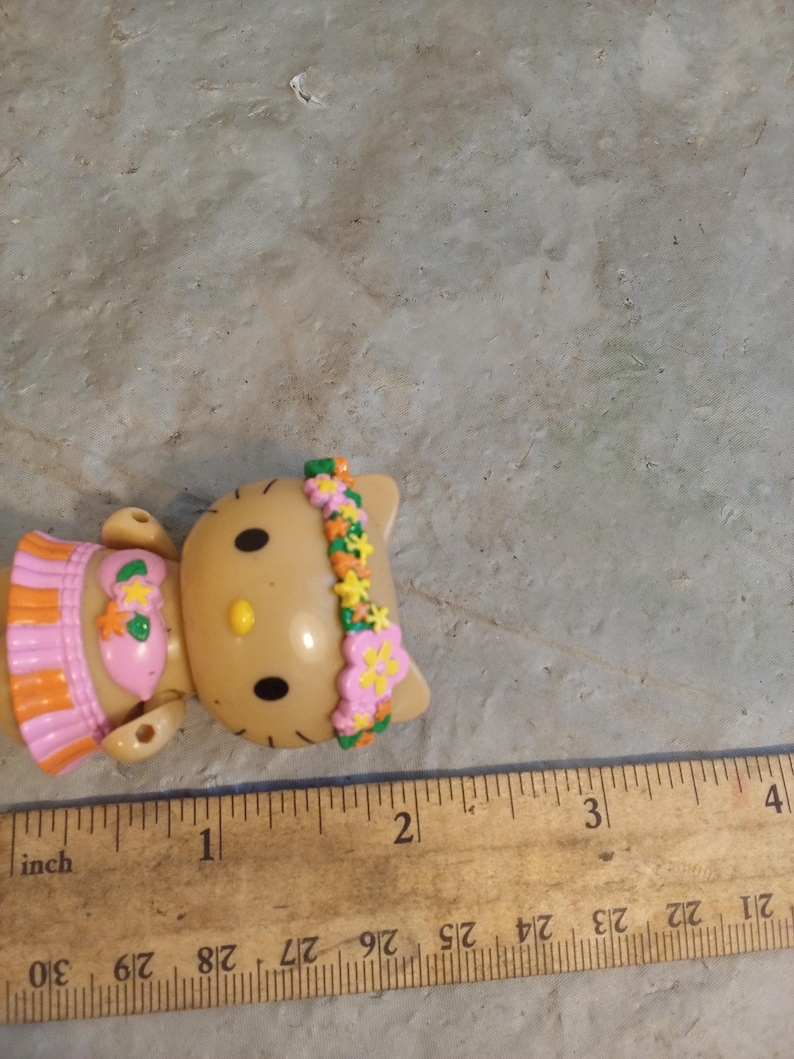 Op de afbeelding: Een bruin plastic beeldje van een kat met zwarte ogen en een gele neus. De kat draagt een roze en oranje outfit en een bloemenhoofdband. Een houten liniaal met inch-metingen ligt naast het beeldje.