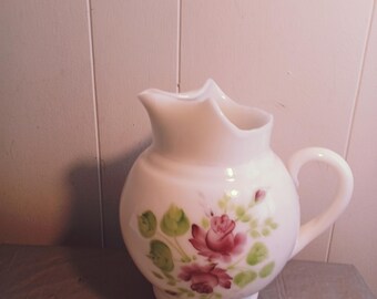 May include: White ceramic jug with a round body, a handle, and a spout. The jug is decorated with pink roses and green leaves. The jug is sitting on a grey surface.