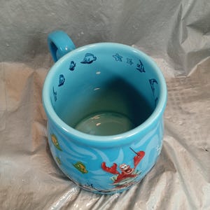 May include: A light blue ceramic mug with a handle, featuring aquatic-themed illustrations. The mug's exterior showcases cartoon images of fish, starfish, and a red crab. The interior is a solid light blue.