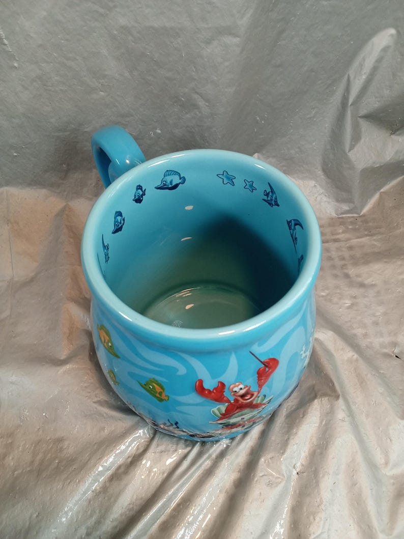 May include: A light blue ceramic mug with a handle, featuring aquatic-themed illustrations. The mug's exterior showcases cartoon images of fish, starfish, and a red crab. The interior is a solid light blue.