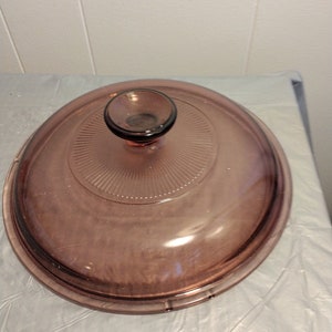 May include: A clear glass lid with a knob on top. The lid is a light purple color and has a ribbed pattern.