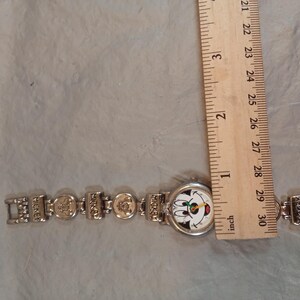 May include: A silver-tone wristwatch featuring a round face with a Mickey Mouse graphic, positioned next to a wooden ruler showing a diameter of approximately 2.5 cm. The metal link band includes rectangular segments embossed with the name Mickey.
