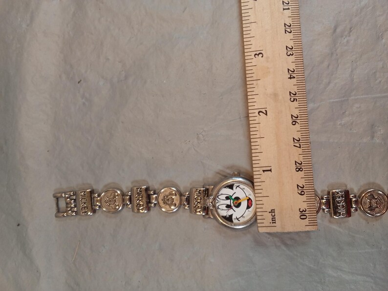 May include: A silver-tone wristwatch featuring a round face with a Mickey Mouse graphic, positioned next to a wooden ruler showing a diameter of approximately 2.5 cm. The metal link band includes rectangular segments embossed with the name Mickey.