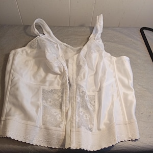 Könnte beinhalten: Weißes Bustier aus satinartigem Material mit Spitzendetails auf den Frontpanels und Trägern. Das Bustier hat einen gewellten Saum und einen Frontverschluss. Das Kleidungsstück ist mit einer strukturierten Silhouette entworfen und ist wahrscheinlich Vintage oder Vintage-inspiriert.